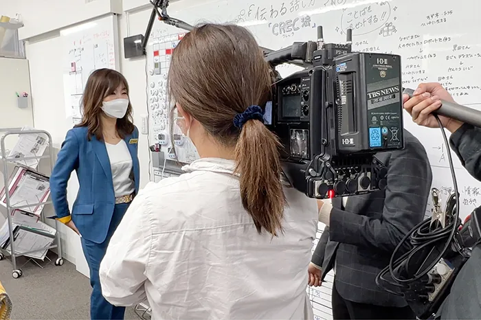 NEWS23 の取材。テレビカメラの前でホワイトボードを背に話すスタッフ